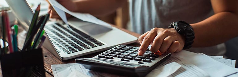 Pessoa utilizando uma calculadora e um computador, representando a análise de relatórios gerenciais para a clínica ou consultório.